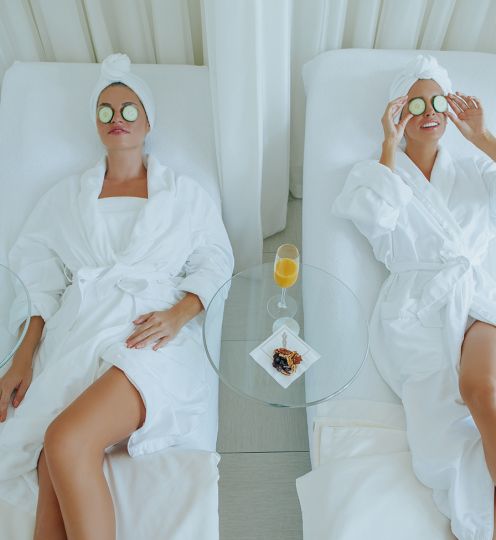 Two women in white robes and towels on their heads lay on chairs with cucumbers on their eyes