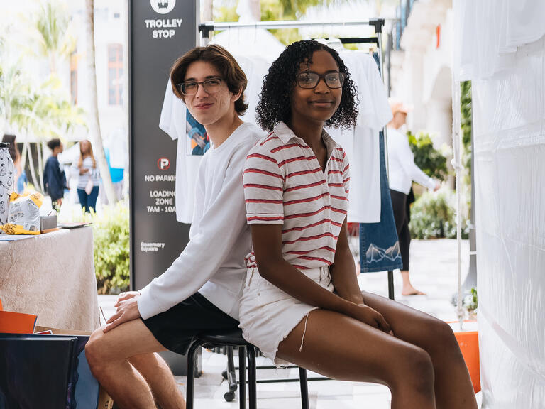 Two young adults sitting on a stool behind a merch table