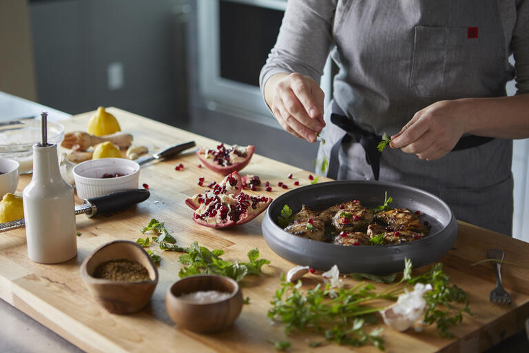 chef completing preparation of meal on circular plate on wooden block on counter surrounded by small dishes of spices and pomegranates
