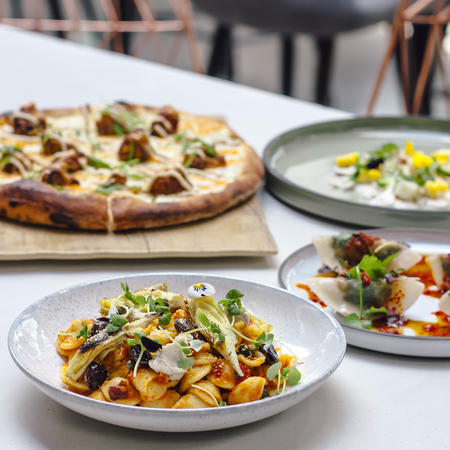 pizza, pasta, and appetizers laid out on white table with chairs on background